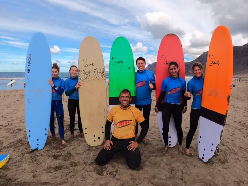 A group of beginner and refresher surfers with their surfboards and the trainer in front of a beach and mountain panorama