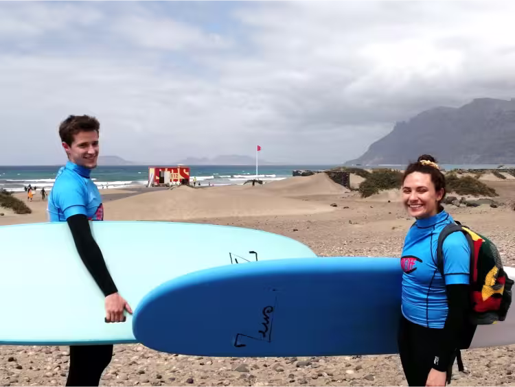 2 persons equiped with wetsuit, take-off surfschool lycra, and surfboards, carrying backpacks n their backs, smiling towards the camera in front of a beach panorama, ready to start their surflesson
