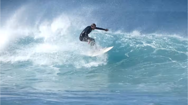 A surfer riding the shoulder of a breaking wave, escaping the white foam, while a strong offshore wind turns the crest into a fine misty spray.