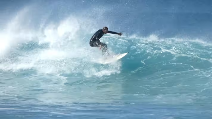 A surfer riding the shoulder of a breaking wave, escaping the white foam, while a strong offshore wind turns the crest into a fine misty spray.